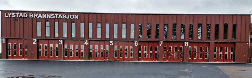 Langt toetasjers bygg i rustrød farge. 9 porter i første etasje. Lystad brannstasjon skrevet øverst til venstre.